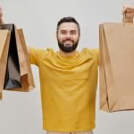 Un homme heureux en chemise jaune tenant plusieurs sacs de courses, exprimant la joie du shopping.
